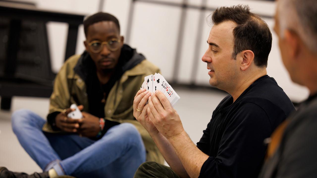 Steve Cuiffo穿着海军蓝衬衫，手里拿着一副扑克牌在演示. 一个学生坐在地板上，专心地观察着. 设置是非正式的和教育的，暗示动手学习的经验.